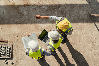 Three people wearing construction site clothing and helmets looking at plans and a laptop on a construction site.