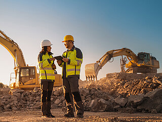 Woman and man wearing construction site clothing on a construction site with excavators in the background.
