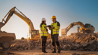Woman and man wearing construction site clothing on a construction site with excavators in the background.