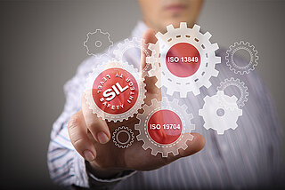 Gears with the inscription “SIL”, “ISO 13849” and “ISO 19704” with a man in the background pointing to the gears